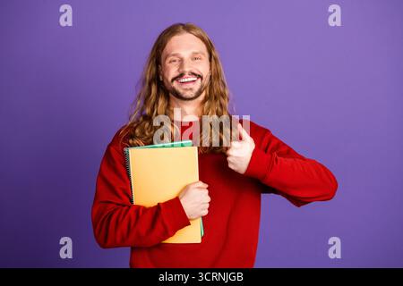 Glücklicher junger Mann mit langen Haaren, der Notizbücher hält und die Daumen vor violettem Hintergrund hochhält Stockfoto