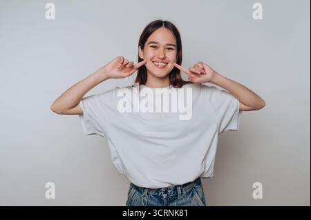 Eine junge Frau in lässiger Kleidung lächelt und zeigt auf ihre Wangen. Stockfoto