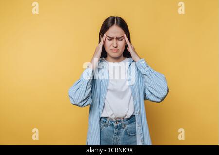 Eine junge Frau mit Kopfschmerzen, die auf gelbem Hintergrund steht und legere Kleidung trägt. Stockfoto