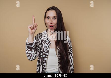 Eine überraschte Frau mit langen Haaren hebt ihren Zeigefinger und trägt ein Hemd mit Zebramuster vor beigem Hintergrund. Stockfoto