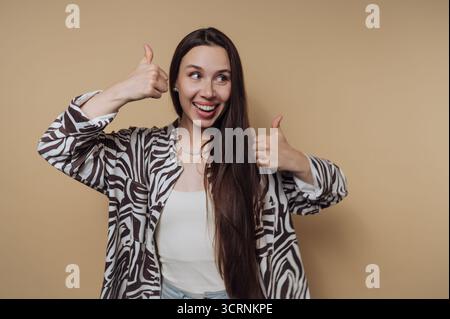 Eine fröhliche Frau mit langen Haaren zeigt sich vor beigefarbenem Hintergrund doppelt hoch. Stockfoto
