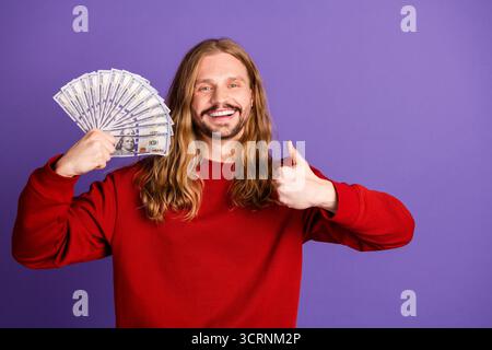 Glücklicher, gutaussehender junger Mann mit langen blonden Haaren, der Bargeld hält und die Daumen in violettem Hintergrund hochgibt Stockfoto