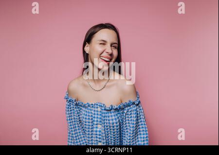 Junge Frau in blau kariertem Off-Schulter-Top lacht vor rosa Hintergrund. Stockfoto