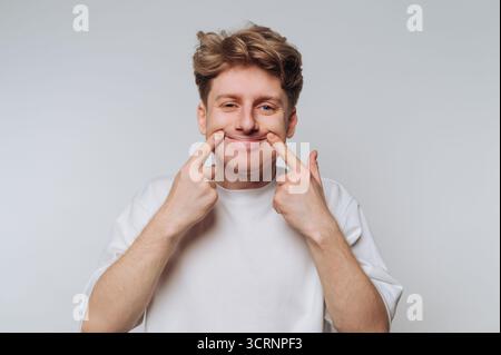 Junger Mann in weißem Hemd, der lächelt und seine Wangen zieht. Stockfoto