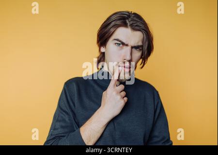 Porträt des jungen Mannes posiert nachdenklich mit einem Finger auf den Lippen und hält einen Moment der Besinnung auf gelbem Hintergrund fest Stockfoto