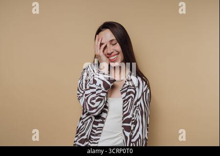Eine Frau steht vor einem warmen Hintergrund und lächelt hell, während sie ihr Gesicht spielerisch mit einer Hand bedeckt und ihre fröhliche Persönlichkeit zum Ausdruck bringt Stockfoto