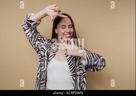 Eine Frau zeigt ihre verspielte Seite, indem sie ihr Gesicht mit den Händen umrahmt, ein gemustertes Hemd trägt und vor einem weichen, neutralen Hintergrund fröhlich lächelt Stockfoto