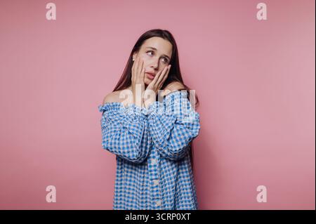 Eine Frau mit langen Haaren zeigt einen besorgten Ausdruck, während sie ihre Hände auf ihr Gesicht legt. Sie trägt ein blau-weiß kariertes Off-Shoulder-Top. Stockfoto