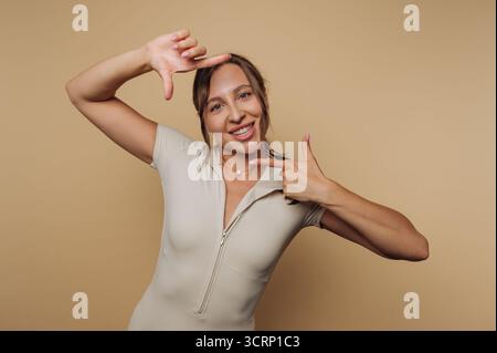 A woman is playfully framing her face with her hands, smiling at the camera. The background is light beige, creating a warm, inviting atmosphere. Stockfoto