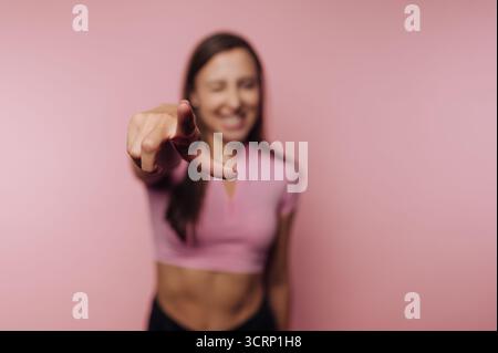 Eine junge Frau in einem rosa Oberteil steht mit einem fröhlichen Ausdruck, der mit dem Finger spielerisch auf den Betrachter zeigt, vor einem sanften rosa Hintergrund und strahlt ein f aus Stockfoto
