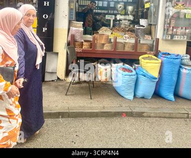 Zwei Frauen laufen an einem typischen Marktladen vorbei, der Gewürze, Kaffee, Tee und andere Speisen in Tanger, Marokko, verkauft. Sie tragen typisch muslimische Kleidung und einen Hijab. Das Essen wird auf traditionelle und offene Weise präsentiert. Stockfoto