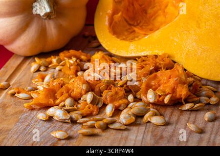 Detaillierte Nahaufnahme von rohen Butternusskürbis-Samen und Orangenschnitzel auf einem hölzernen Schneidebrett, mit geschnittenen Kürbishälften im Hintergrund. Natürliches Herbstgemüse Stockfoto