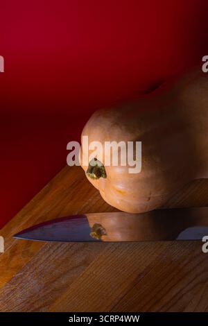 Nahaufnahme eines ganzen Butternusskürbis auf einem hölzernen Schneidebrett mit einem großen Küchenmesser, das das Gemüse reflektiert. Dramatische Beleuchtung mit Tiefenbeleuchtung Stockfoto