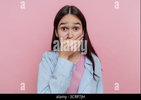 Überraschte junge Frau, die den Mund mit der Hand vor rosafarbenem Hintergrund bedeckt. Stockfoto