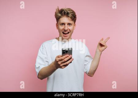 Aufgeregter junger Mann mit Smartphone, das auf etwas zeigt. Stockfoto