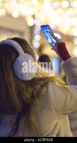 Eine junge Frau, die Ohrenschützer trägt, hebt ihr Smartphone an, um ein Foto von glühenden festlichen Lichtern in der Nacht zu machen, um einen Moment der städtischen Freude und der Jahreszeit festzuhalten Stockfoto