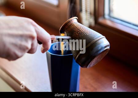 Kaffee aus einer Cezve wird in einen hohen, dunklen Keramikbecher auf einer Fensterbank gegossen. Dampf steigt auf, während Tageslicht die Atmosphäre des Komforts verbessert. Stockfoto