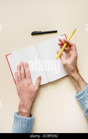 Männliche Hände (Rechtshänder) schreiben mit Bleistift auf eine leere Seite eines offenen Notizbuchs. Draufsicht auf neutralem Hintergrund. Ideal für Journale und Studien. Stockfoto