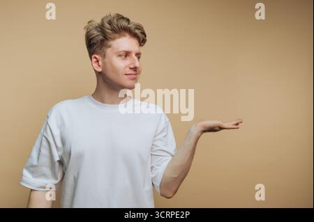 In einem neutralen Innenraum steht ein junger Mann mit einem leichten Lächeln und streckt seine rechte Hand nach vorne und lädt zur Verlobung ein. Er ist entspannt Stockfoto