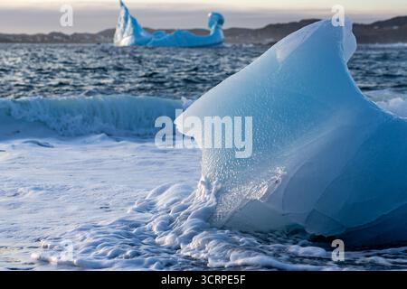 Eine Nahaufnahme eines kleinen Eisbergs an einem Strand, mit Wellen, die um ihn herum wehten. Im Hintergrund gibt es größere Eisberge, die im Ozean schwimmen, und eine Stockfoto