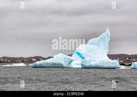 Ein großer Eisberg schwimmt im Meer in der Nähe einer kleinen Stadt an einem bewölkten Tag. Stockfoto