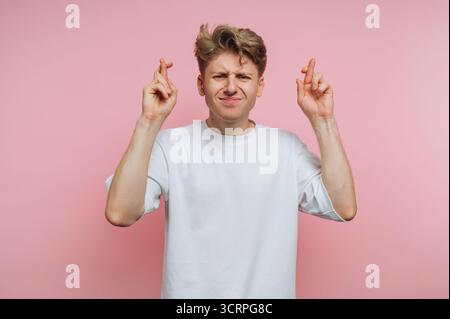 Porträt eines jungen Mannes, der vor rosa Hintergrund die Finger kreuzt und Hoffnung und Positivität auf rosa Hintergrund symbolisiert Stockfoto