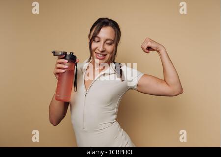 Porträt einer fitten jungen Frau spannt ihre Muskeln, während sie eine Wasserflasche hält, und verkörpert Kraft und Begeisterung für Fitness auf beigefarbenem Hintergrund Stockfoto