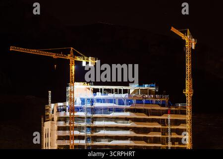 Ein eindrucksvolles Nachtfoto mit zwei hohen Baukränen, die vor einer dunklen, bergigen Kulisse in Benidorm beleuchtet werden. Die Kräne stehen über einem Teil Stockfoto