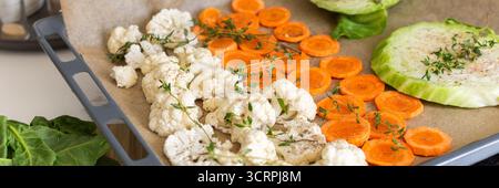 Blumenkohl, Karotten und Kohl werden in Scheiben geschnitten und auf einem mit Pergamentpapier ausgelegten Tablett angeordnet, fertig zum Rösten mit Kräutern in einem warmen Küchenset Stockfoto
