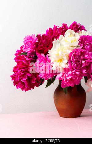 Ein lebhaftes Arrangement aus rosa und weißen Pfingstrosen füllt eine Keramikvase, die auf einer weichen rosafarbenen Tischdecke platziert wird. Die Blumen verleihen jedem Raum eine fröhliche Note. Stockfoto