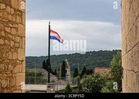 Eine einsame kroatische Flagge schwingt auf einem hohen Pfosten zwischen Steinmauern und überblickt ein ruhiges Dorf mit Terrakottadächern und üppigen Hügeln in der Ferne. Kal Stockfoto