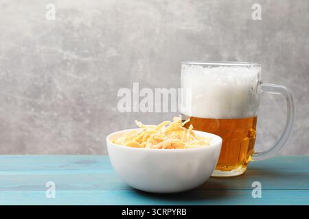Getrocknete Tintenfischspäne und Glasbecher Bier auf hellblauem Holztisch vor grauem Hintergrund, Nahaufnahme. Leerzeichen für Text Stockfoto