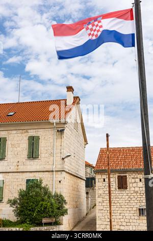 Eine kroatische Flagge fahnt über einem Steinhaus mit grünen Fensterläden und einem rot gekachelten Dach vor einem hellblauen Himmel. Eine heitere, ländliche Szene, die so toll ist Stockfoto