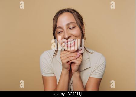 Eine fröhliche junge Frau lächelt breit mit den Händen unter ihrem Kinn. Der warme Hintergrund ergänzt ihr positives Verhalten und ihren lebendigen Ausdruck Stockfoto