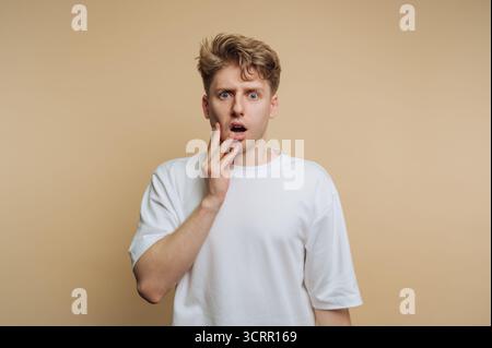 Ein junger Mann mit lockigen blonden Haaren sieht schockiert aus und berührt sein Gesicht mit einer Hand. Der schlichte beige Hintergrund unterstreicht sein überraschendes Verhalten in A Stockfoto