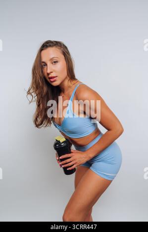 Eine Frau in blauer Sportkleidung steht selbstbewusst in einem Studio und hält eine Shaker-Flasche in der Hand. Ihr Haar fließt nach unten, während sie eine dynamische Pose schlägt. Stockfoto