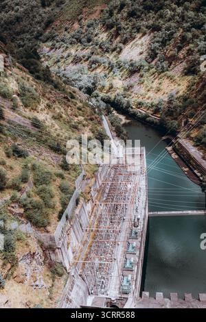 Aus der Vogelperspektive eines Wasserkraftwerks, das in einen steilen grünen Canyon gebaut wurde, mit Übertragungsleitungen, Betondamm und Fluss, der darunter fließt, symbolisiert Stockfoto