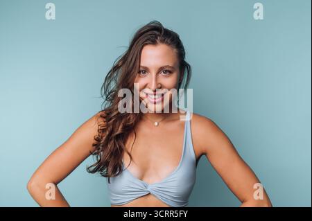 Eine Frau steht mit einem fröhlichen Ausdruck und trägt ein hellblaues Top. Ihr langes, gewelltes Haar fließt frei, während sie eine selbstbewusste Pose gegen ein weiches P anschlägt Stockfoto
