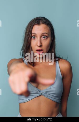 Eine Frau mit langen Haaren drückt Schock und Unglauben aus, während sie nach vorne zeigt. Sie steht vor einer hellblauen Wand und trägt ein lässiges Outfit. Der geldautomat Stockfoto