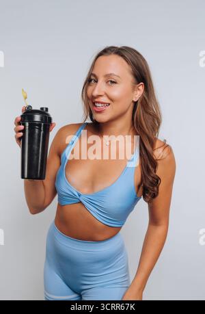 Eine junge Frau posiert selbstbewusst in blauer Trainingskleidung und hält eine schwarze Shaker-Flasche. Sie hat lange Haare und ein helles Lächeln, zeigt einen passenden lifestyl Stockfoto