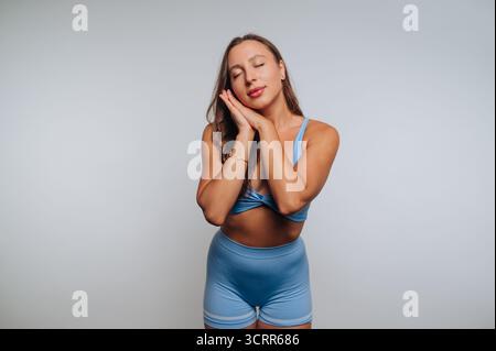 Eine Frau in hellblauer Sportkleidung steht mit geschlossenen Augen und Händen nahe ihrem Gesicht und vermittelt eine friedliche Atmosphäre in einer einfachen Innenumgebung. Stockfoto