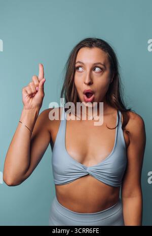 Eine Frau in blauer Activewear hebt ihren Zeigefinger mit einem überraschenden Ausdruck, als ob sie eine plötzliche Idee oder Erkenntnis teilt. Der Hintergrund ist Stockfoto