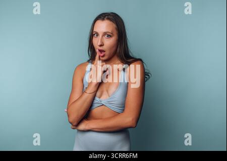 Eine Frau steht vor einem hellblauen Hintergrund und trägt ein modisches Outfit. Sie sieht überrascht aus und legt ihren Finger spielerisch auf die Lippen. Stockfoto