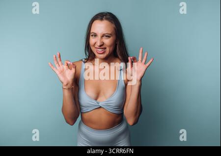 Ein junger Mensch steht vor einem hellblauen Hintergrund und trägt ein anliegendes Outfit. Sie machen spielerische Gesichtsausdrücke und Handgesten, Showcasi Stockfoto