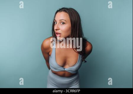 Eine junge Frau zeigt eine verspielte Pose, während sie ein hellblaues Sportoutfit trägt. Ihr Ausdruck vermittelt Selbstvertrauen vor einem passenden blauen Hintergrund. Stockfoto
