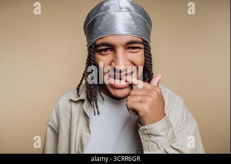Ein junger Mann mit geflochtenen Haaren und silbernem Kopftuch lächelt und berührt spielerisch seine Zähne. Er posiert vor einem neutralen Hintergrund und zeigt eine lustige m Stockfoto