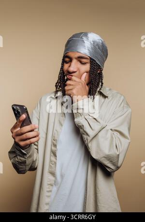 Ein junger Mann, der eine silberne Kopfhülle und ein leichtes Hemd trägt, scheint tief in Gedanken zu sein, als er sein Smartphone überprüft. Der neutrale Hintergrund verleiht dem HIS mehr Aufmerksamkeit Stockfoto