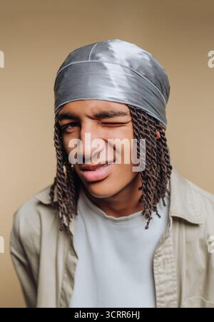 Ein junger Mann zwinkert spielerisch, während er ein Kopftuch trägt. Seine stylischen Dreadlocks umrahmen sein Gesicht, während er eine selbstbewusste Pose gegen einen weichen Neutren schlägt Stockfoto