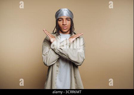 Eine junge Person steht selbstbewusst mit gekreuzten Armen, trägt einen glänzenden Kopfschutz und ein gedämpftes Outfit. Die neutrale Kulisse verstärkt die markante Gestu Stockfoto