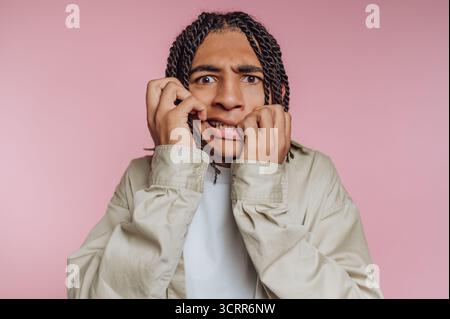Ein junger Mann mit geflochtenen Haaren zeigt einen schockierten Ausdruck, indem er seine Wangen greift. Der Hintergrund ist in zartem Rosa gehalten und sorgt für eine verspielte und lebendige Atmosphäre Stockfoto